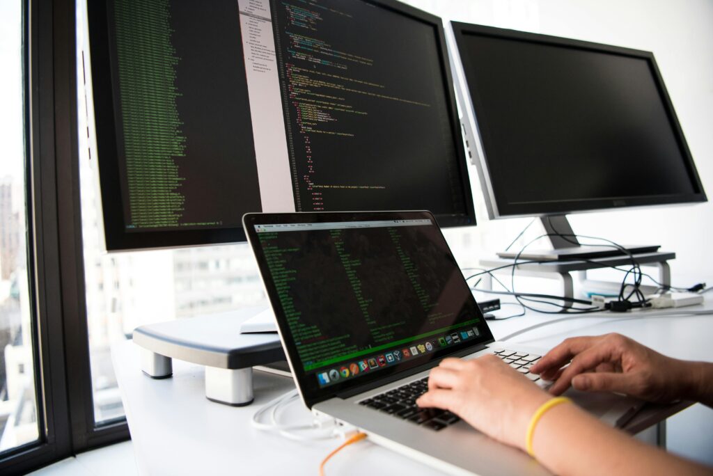 pexels photo 1181675 1181675 A developer writes code on a laptop in front of multiple monitors in an office setting.